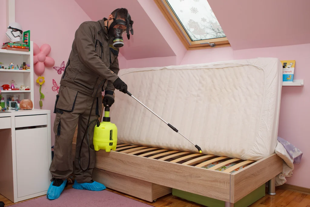 A masked professional sprays pest control under a raised mattress in a child's bedroom, emphasizing safety and hygiene. Venus Pest solution in Lahore.