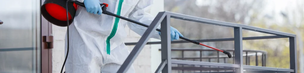 A person in protective gear applies a cleaning solution to a metal railing using a spray device on a bright day, ensuring pest control for commercial buildings.