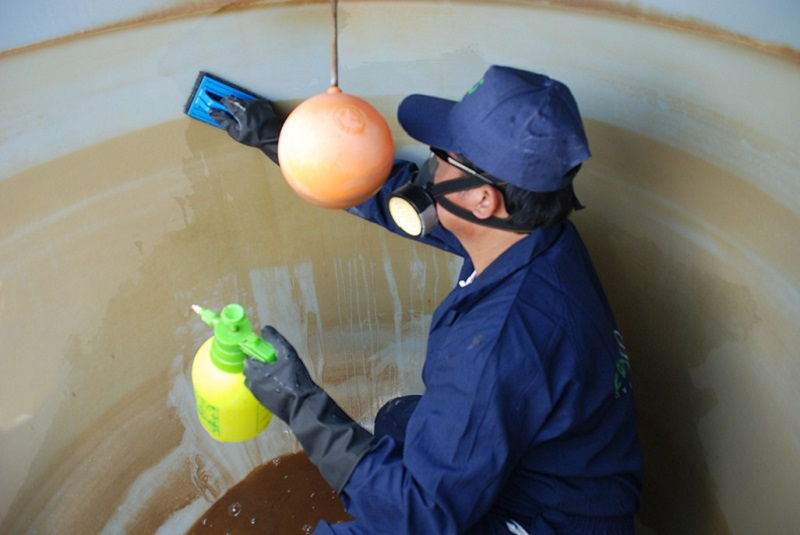 A worker in protective gear cleans the interior of a large tank, using a spray bottle and sponge to remove residues during Water Tank Cleaning.