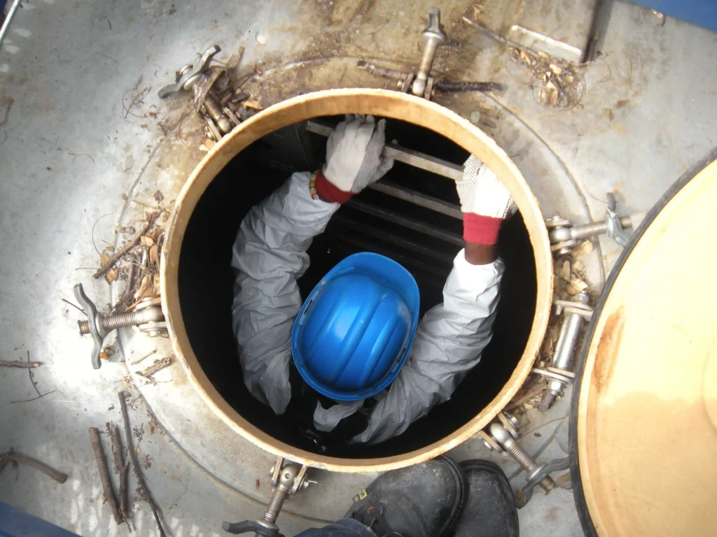 A worker in protective gear and a blue helmet descends into a large circular tank or pit, gripping metal rungs for support during Water Tank Cleaning.