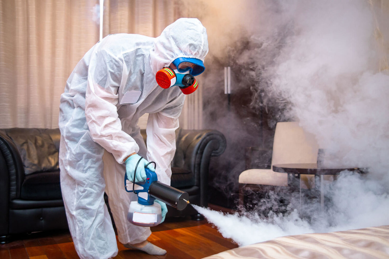 A person in a protective suit and mask sprays disinfectant during a fumigation service in a smoke-filled room, ensuring cleanliness and safety.