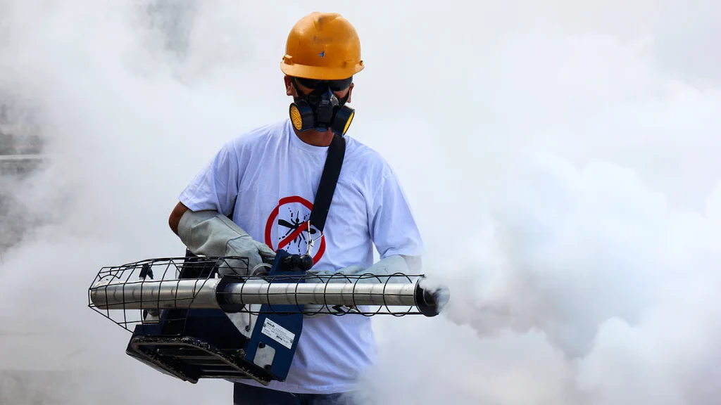A worker in a yellow hard hat and protective gear operates a fogging machine, releasing white smoke as part of a mosquitoes spray treatment for effective pest control.