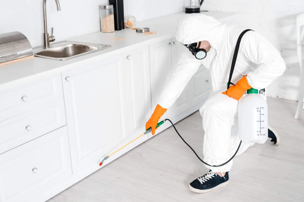 Person in protective gear performing rodent control in white kitchen, spraying pesticide at cabinet base while wearing hazmat suit, gas mask, and orange gloves.