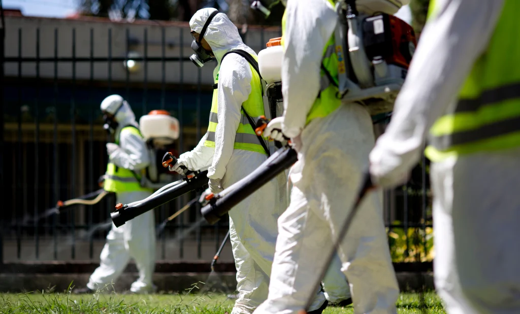 Workers in protective gear perform pest control in Lahore, spraying pesticide in a green area using backpack sprayers and long-handled tools.