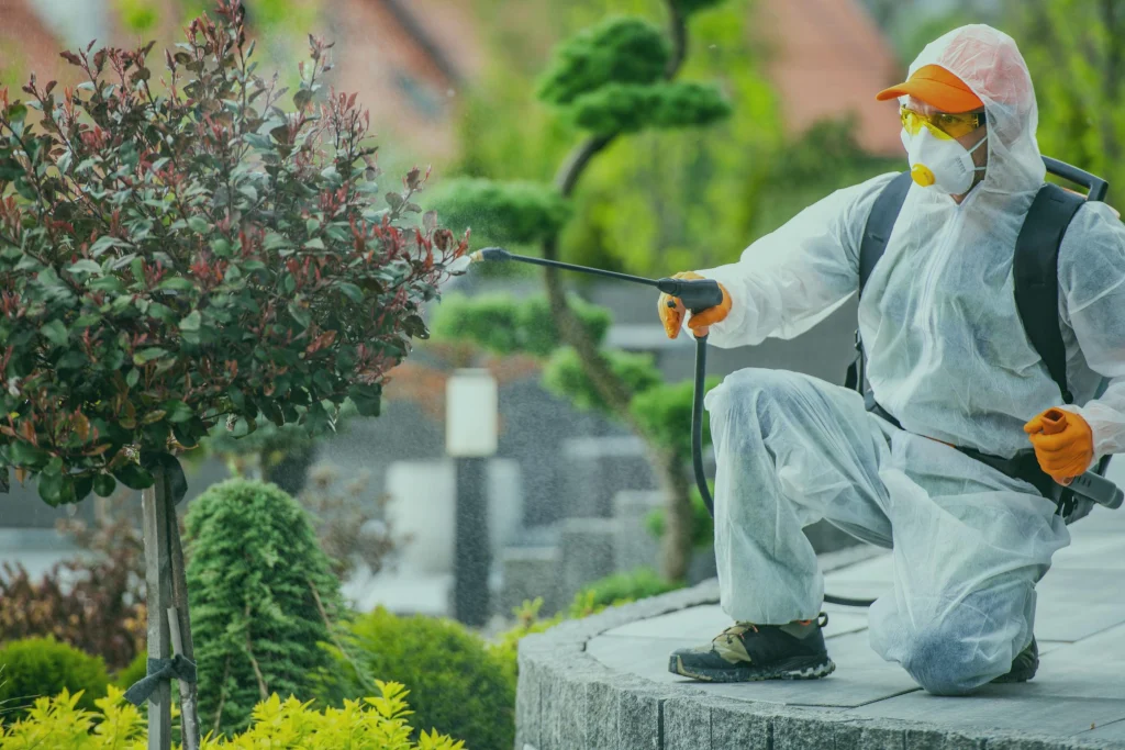 A pest control expert in protective gear sprays a plant with pesticide in a landscaped garden, representing pest control in Lahore.