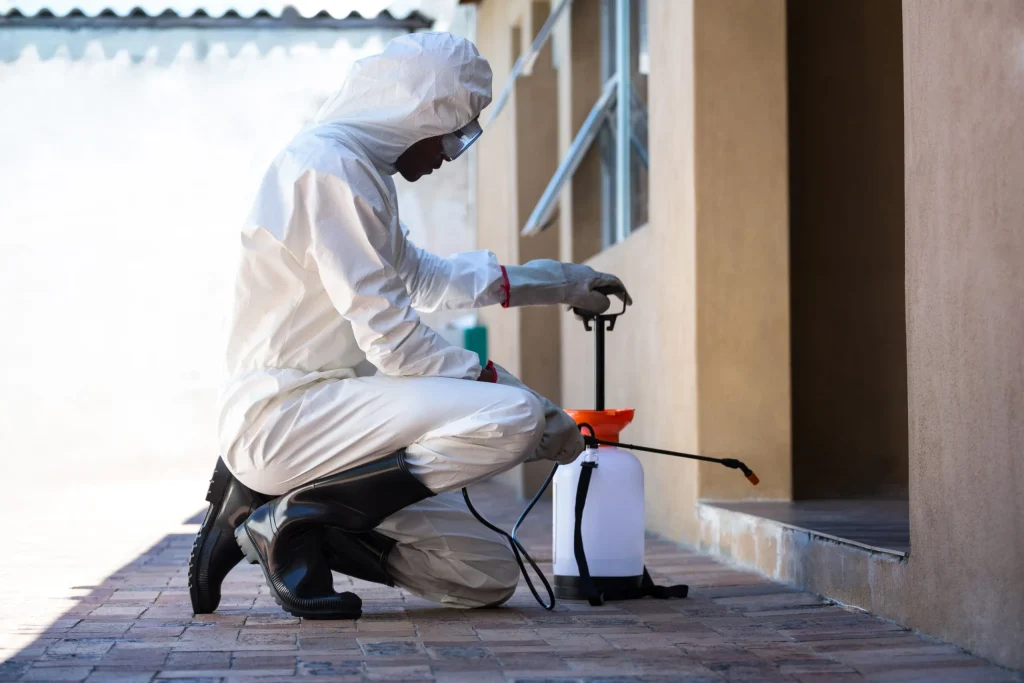 A person in a white protective suit kneels to operate a sprayer for pesticide application near a building's entrance — part of effective Rainy Season Pest Solutions in action.