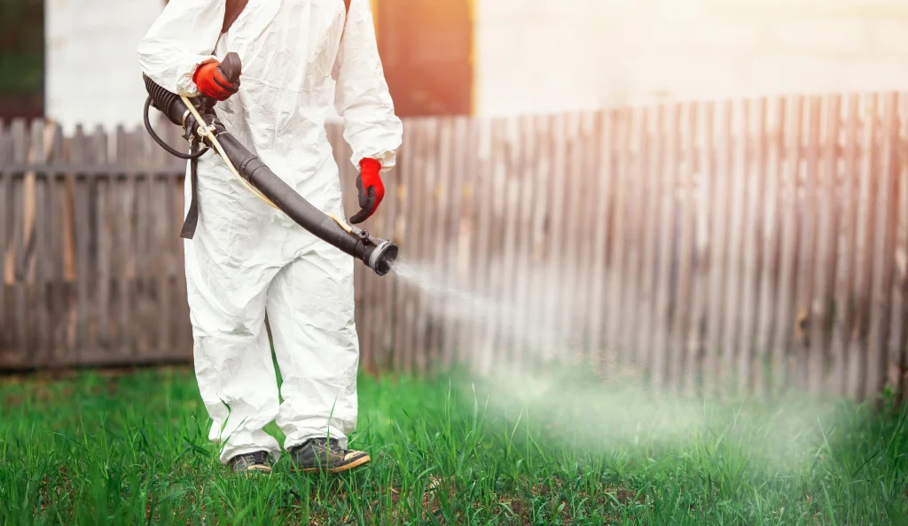 A person in a white protective suit sprays mosquitoes spray on green grass using a long nozzle, with a wooden fence in the background.