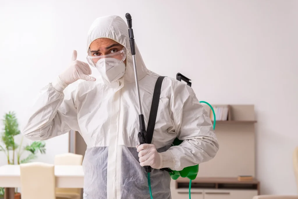 A pest control professional in full protective gear, including a face mask and gloves, stands in a bright room holding equipment. They gesture “call me”, offering expert pest control in Sialkot.